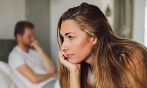 woman sits on bed thinking while man sitting hand in face in corner feeling unhappy