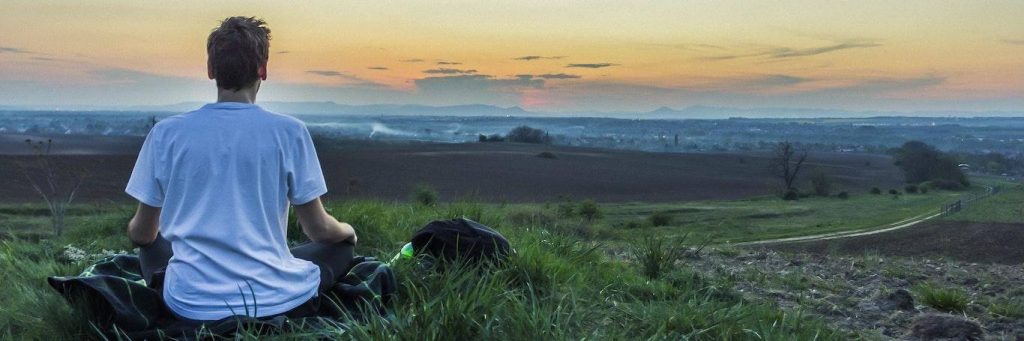 man sits on green grass facing backward meditating in sunset dark sky