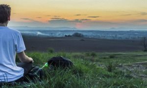 man sits on green grass facing backward meditating in sunset dark sky