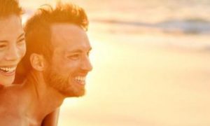 man carries girlfriend on back walking on beach in sunny sky