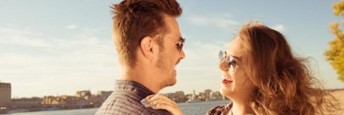 couple wearing sunglasses stands near blue ocean laughing