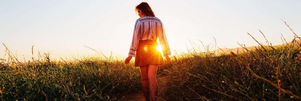 woman facing backward walks along footpath on field in beautiful sunny sky