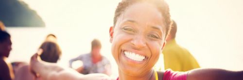 short hair woman happily smiles taking selfie on busy beach in sunny sky