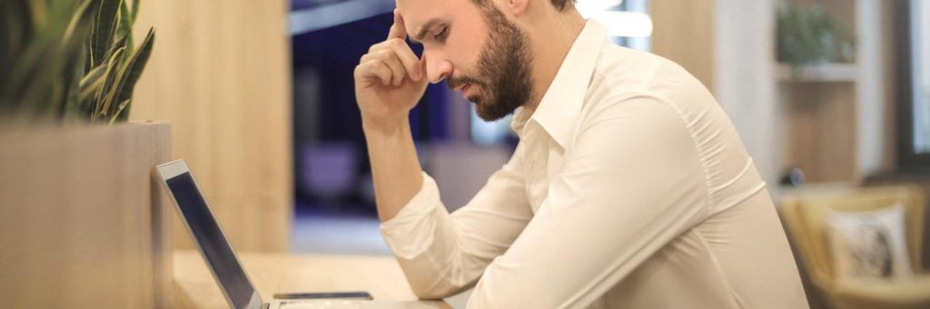 man sits in library hand in forehead thinking looking at laptop