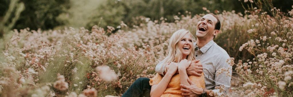 couple sits on field laughing while man hugging partner from behind