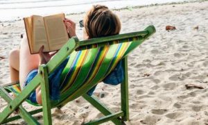 woman wearing sunglasses sits on chair reading book on beach