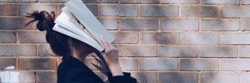 woman holds book covering face stands beside stone wall