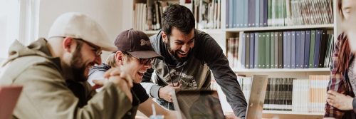 three men sits in library excitedly laughing while using laptop beside bookshelf