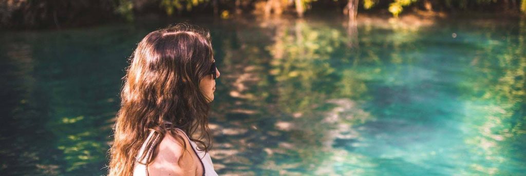 woman wearing sunglasses stands beside blue water lake thinking