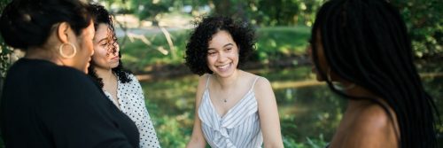 four young women standing in park beside lake speaking laughing