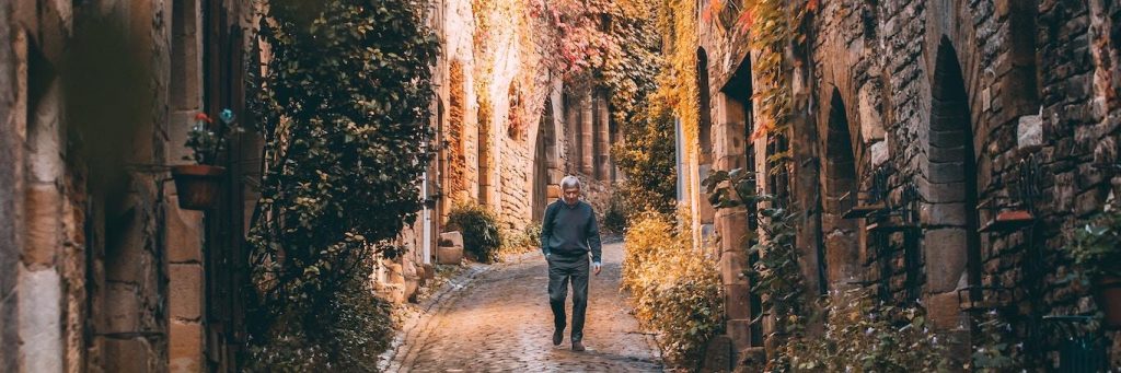 elderly man hand in pocket walks alone along footpath between old buildings