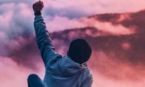 man sits raising hand looking mountain red blue cloudy sky