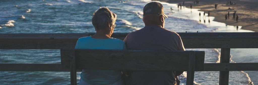 elderly couple sits on bench near beach facing backward enjoying cool weather watching people swimming