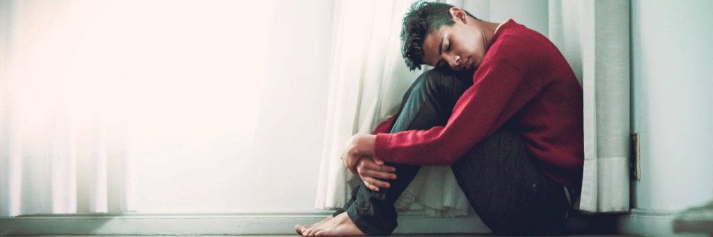 man sits bend forward face on knees sleeping next to white window curtain