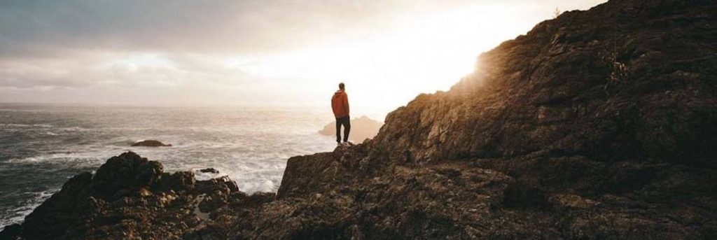 man stands hands in pocket on rock beside ocean looking at sunny sky
