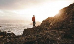 man stands hands in pocket on rock beside ocean looking at sunny sky