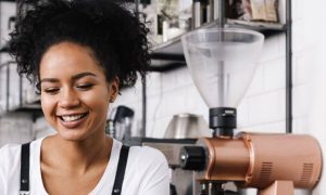 curly black hair woman sits in coffee shop beside coffee machine black shelf happily smile