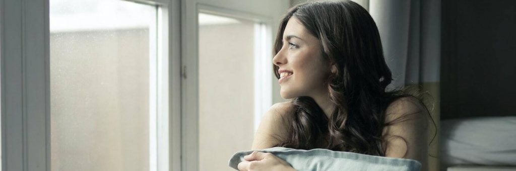 young woman sits beside window smiling hugging white pillow