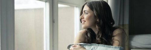 young woman sits beside window smiling hugging white pillow