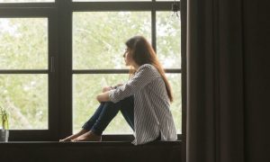 woman sits alone on bench beside window curtain looking outside