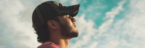 man eyes closed wears black cap meditating in blue cloudy sky