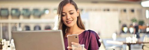 woman sits in coffee shop using mobile phone beside laptop