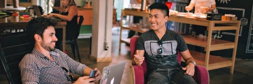 two men sits in busy coffee shop happily talking smiling discussing work
