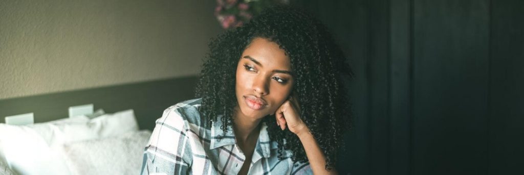 curly black hair woman sits hand on cheek in bedroom thinking