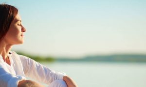 woman eyes closed sits beside lake breathing focusing on inner peace