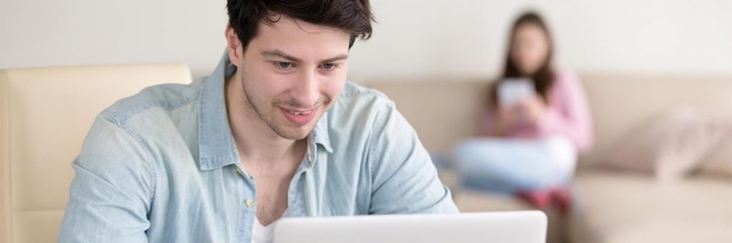 man smiles studying laptop while woman sitting using mobile phone on white couch