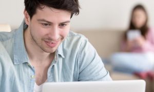 man smiles studying laptop while woman sitting using mobile phone on white couch