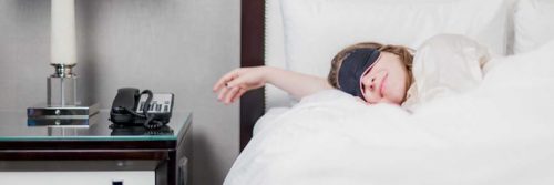 woman wearing mask covering eyes sleep on white bed set beside side table lamp telephone
