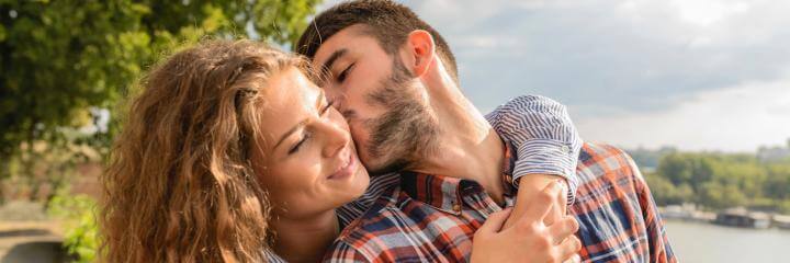man kisses girlfriend on cheek while woman hugging from behind