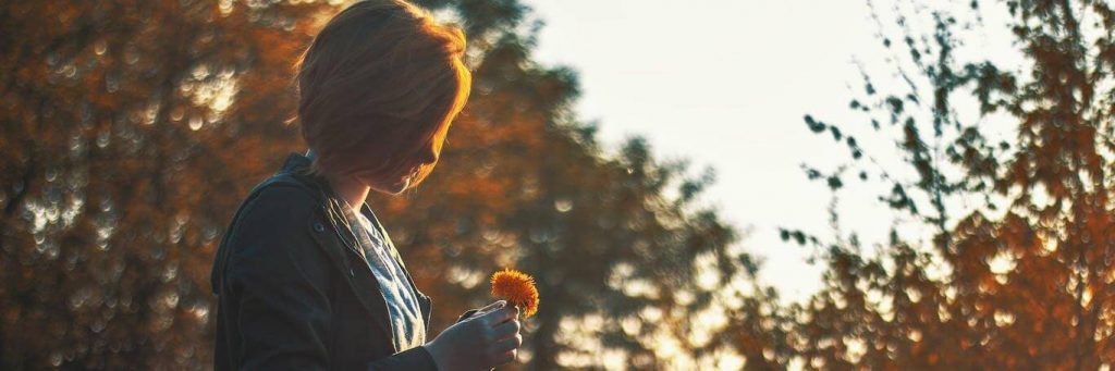 short hair woman holds small orange flower standing in forest in sunny sky