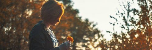 short hair woman holds small orange flower standing in forest in sunny sky