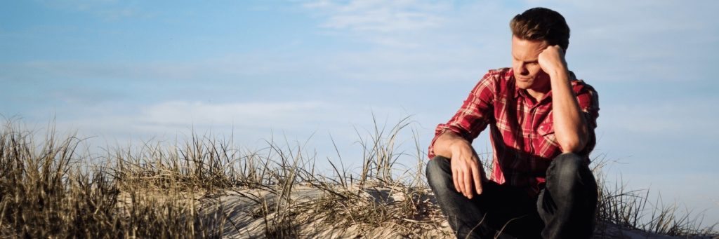 man sits alone on rock hand on head sadly thinking in blue partially clear sky