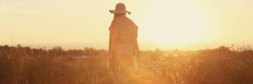 woman wearing hat facing backward walk alone on field in sunny sky