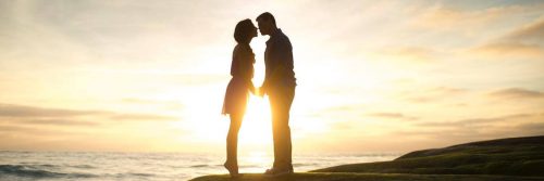 couple stands on rock beside ocean kissing in beautiful sunset