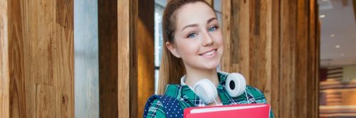 girl teenager wearing headphone stands in library holding red cover document smiling