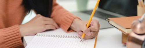 woman hand holding pencil writing on white notebook beside laptop