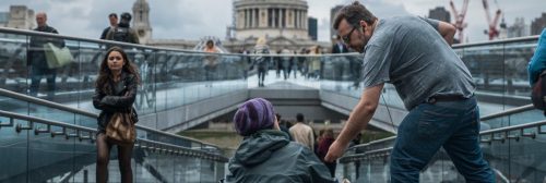 man wearing hat sits asking receiving support from man walking by in busy crowded building