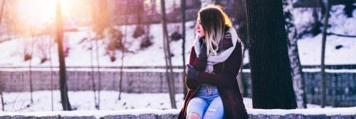woman wearing winter coat sits on fence in snowy forest in shining sun