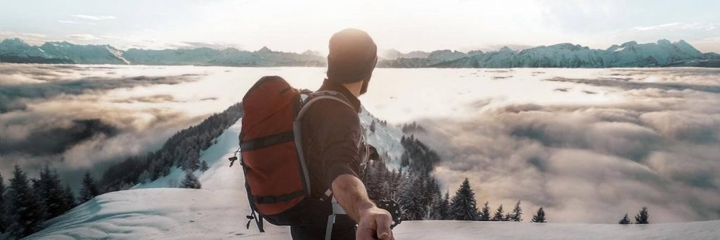 man wearing back bag stands on snowy mountain holding selfie stick looking at mountain in nice sunny weather