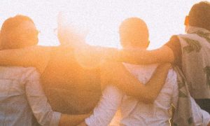 group of close friends stands facing backward hugging each other looking at sunny sky