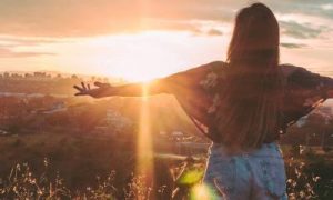 woman facing backward stands on field raising hands gratitude life in peaceful environment in sunny sky