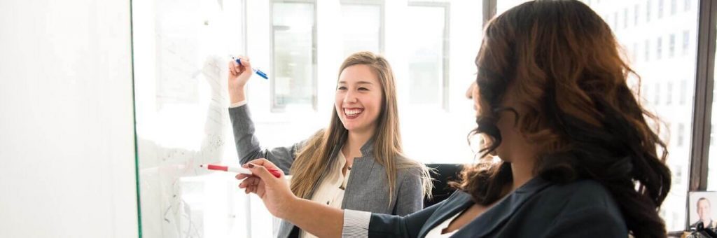 two business women stands in office happily discussing writing on whiteboard