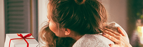 two women hug beside white giftbox wrapped by red banner