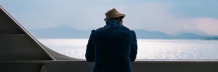 man facing backward stands on boat looking at ocean in blue cloudy sky