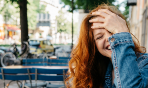 long curly brown hair woman with happy face hand on forehead laughs standing next to restaurant