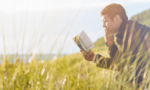man sits on chair in field beside lake focusing on reading book in cloudy sky